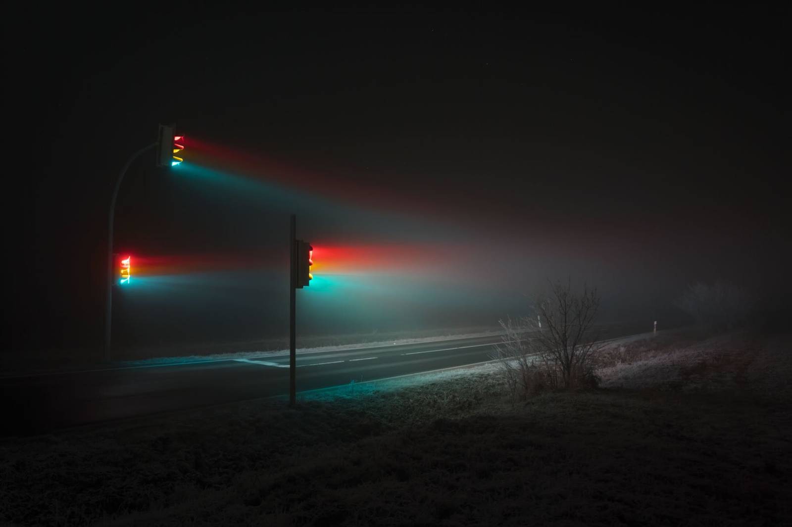 Traffic lights glowing red, yellow, and green at night, with beams of light cutting through dense fog over a deserted road.