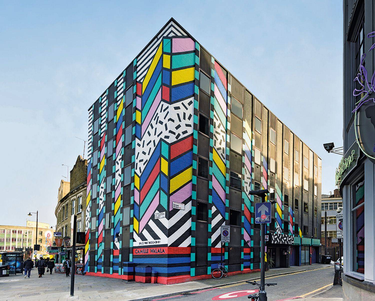Colorful geometric mural by Camille Walala covering a multi-story building on a city street corner, with bold patterns in bright shades and black-and-white stripes.