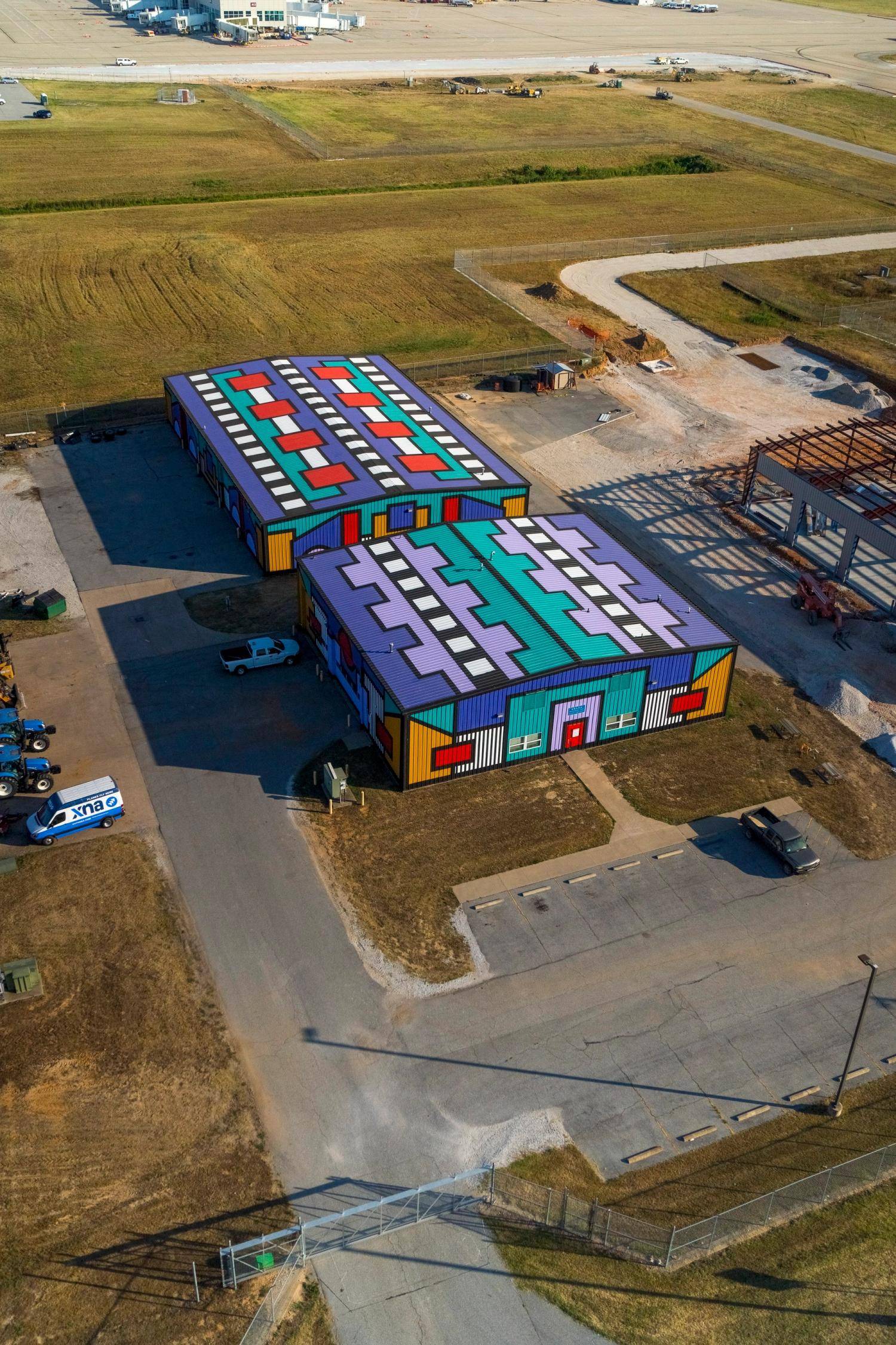 Aerial view of two industrial buildings painted in bold, colorful geometric patterns with graphic designs covering the roofs and walls, located near an airport runway.