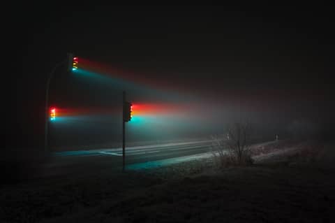 Traffic lights glowing red, yellow, and green at night, with beams of light cutting through dense fog over a deserted road.