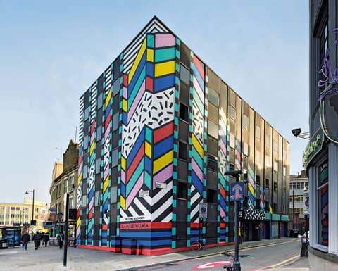 Colorful geometric mural by Camille Walala covering a multi-story building on a city street corner, with bold patterns in bright shades and black-and-white stripes.