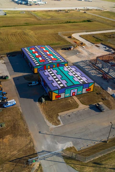 Aerial view of two industrial buildings painted in bold, colorful geometric patterns with graphic designs covering the roofs and walls, located near an airport runway.