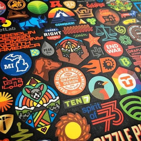 Close-up of a colorful collage of graphic design logos, icons, and badges printed on a textured black surface, featuring bold typography, illustrations, and symbols in various styles.