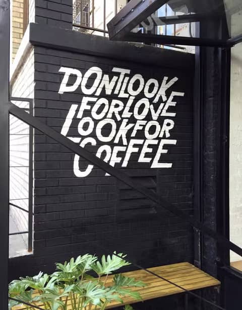 White painted wall mural on a black brick wall that reads “DON’T LOOK FOR LOVE LOOK FOR COFFEE,” located outside a café with glass framing, a small wooden bench, and a leafy green plant in the foreground.